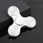 Spinning fidget en métal blanc avec trois bras, conçu pour la détente et la concentration, posé sur une surface noire élégante.
