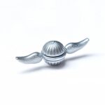 Petite boule en métal argenté avec des ailes, inspirée d’un objet scientifique ou décoratif, posée sur un fond blanc.