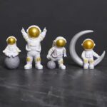 Quatre figurines d'astronautes en décoration, avec des casques dorés et des costumes blancs, accompagnées d'une lune en décoration, parfaites pour un thème spatial ou scientifique.