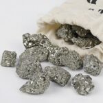 Pépite de pyrite brillante et métallique, provenant d'une collection scientifique, disposée sur une surface blanche avec un sac en toile en arrière-plan.