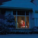 Père Noël vêtu de rouge, regardant par la fenêtre illuminée de la maison enneigée la nuit.