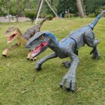 Deux dinosaures T-Rex en peluche géants pour décoration, posés sur une pelouse dans un parc, avec des arbres en arrière-plan.