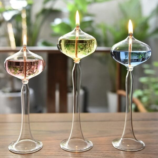 Trois vases en verre transparent avec bougies allumées, contenant des liquides colorés (rose, vert, bleu), sur une surface en bois avec un fond flou de plantes.