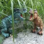 T-Rex en peluche coloré dans un environnement naturel avec herbes et sol sableux.