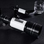 Télescope astronomique noir avec lentille large, posé sur une surface humide, avec un verre d'eau à proximité, illustrant l'observation scientifique et l'astronomie.