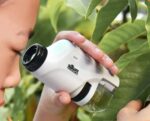 Jeune enfant utilisant un vérificateur de plantes portatif pour examiner les feuilles d’un arbre, dans un environnement naturel verdoyant.