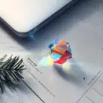 Pendentif en cristal prismatique coloré projetant un arc-en-ciel sur une surface blanche, posé sur un document avec une branche de pin à côté.