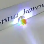Pendentif en verre coloré avec lumière LED intégrée, posé sur une surface blanche, avec un fond de texte partiel "anna karen".