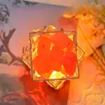 Pendentif en forme de cube avec pierres orange illuminées, encadré par une structure métallique dorée, posé sur une surface en bois avec un fond flou de fleurs et de nature.