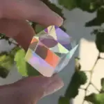 Un cube en cristal irisé avec des reflets multicolores, tenu entre deux doigts, avec un fond de feuilles vertes.