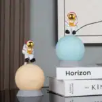 Deux figurines d'astronautes en plastique avec casques dorés, posées sur des sphères lumineuses, décorant un intérieur moderne avec des livres en arrière-plan.