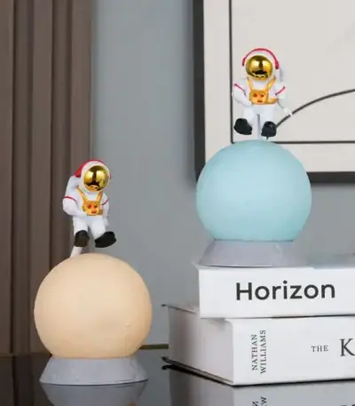 Deux figurines d'astronautes en plastique avec casques dorés, posées sur des sphères lumineuses, décorant un intérieur moderne avec des livres en arrière-plan.