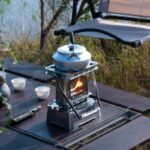 Pique-nique en plein air avec réchaud à gaz et théiÚre en aluminium sur une table en bois, entourée de nature, parfait pour une aventure en camping ou une sortie en plein air.