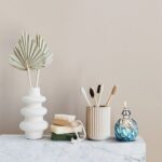 Vase blanc avec feuilles décoratives, brosses à dents dans un pot en céramique, flacon d'huile essentielle bleu, sur une surface en marbre, décoration de salle de bain élégante.