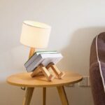 Lampe de table avec livres empilés formant une figure humaine, posée sur une table en bois dans un intérieur moderne.