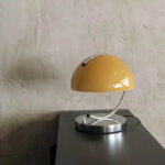 Lampe de bureau vintage en forme de demi-sphère jaune avec bras métallique, posée sur une surface noire contre un mur en plâtre.