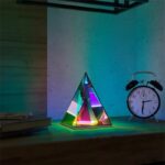 Lampe scientifique en forme de pyramide avec éclairage coloré, accompagnée d'une horloge réveil vintage sur un bureau en bois.