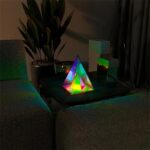 Pyramide en verre colorée avec éclairage LED multicolore, décoratif et moderne, posée sur une table basse dans un salon cosy, entourée de plantes et de meubles modernes.