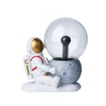 Figurine d'astronaute assis à côté d'une planète en pierre avec une boule de cristal représentant une station spatiale ou un vaisseau, idéale pour la décoration scientifique ou éducative.