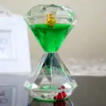 Pendentif en forme de sablier transparent avec liquide vert, décoré de petites figurines colorées à l'intérieur, idéal pour la décoration ou comme porte-clés original.