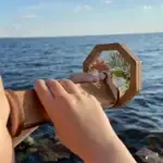 Pendentif en bois avec miroir, décoré de branches de pin et de fleurs, tenu par une main contre un fond d'océan.