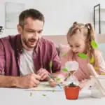 Un père et sa fille réalisent une expérience scientifique à la maison avec un terrarium, un verre à expérience et des feuilles vertes, favorisant l'apprentissage ludique.