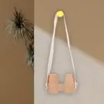 Lunette de vision en bois suspendue avec bande en tissu blanc, fixée au mur avec un bouton jaune, décoration minimaliste et naturelle.