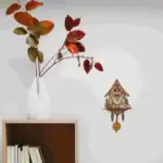 Vase blanc avec branches aux feuilles rouges et vertes, décoratif, avec horloge de style chalet en bois suspendue au mur.