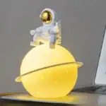 Vaisseau spatial décoratif avec astronaute en costume blanc et casque doré, posé sur une planète lumineuse jaune, idéal pour la décoration scientifique ou thématique.