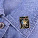 Pince à cravate en métal avec motif spatial et date 1961, fixée sur une chemise en denim bleu clair, ajoutant une touche de style vintage et scientifique à la tenue.