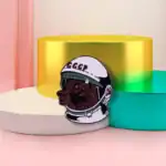 Pendentif en forme de tête de chien portant un casque spatial avec inscription "CCCP", posé sur des supports colorés pastel.