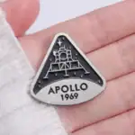 Badge en forme de triangle représentant le module lunaire Apollo 1969, avec détails en relief et inscription "APOLLO 1969", idéal pour collectionneurs et passionnés d'espace.
