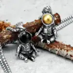 Pendentifs astronautes en métal avec boules de couleur dorée et noire, suspendus à une chaîne, inspirés de l'espace et de l'exploration spatiale.