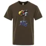 T-shirt marron avec un motif d'astronaute flottant portant un ballon formé de plusieurs planètes colorées, créant une scène spatiale ludique et imaginative.