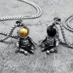 Pendentifs astronautes en métal avec sphères dorée et noire, suspendus à des chaînes, conçus comme des bijoux originaux et modernes inspirés de l'espace.