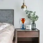 Vase en verre orange et bleu sur une table de nuit moderne avec plante verte et lampe suspendue dorée.