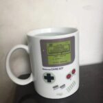 Mug en forme de console Game Boy avec écran affichant "Super Mario Land" et boutons colorés, idéal pour les fans de jeux vidéo rétro.
