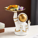 Figurine d'astronaute en céramique blanche avec casque doré, tenant un plateau rempli de bonbons colorés, décoration originale pour un espace thématique ou une collection d'objets scientifiques.