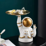 Figurine d'astronaute en céramique blanche et or, tenant un plateau avec des accessoires scientifiques, décor de style cosmique pour collection ou décoration.