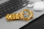 Montre mécanique dorée avec mouvement visible, posée sur un ordinateur portable noir avec des écouteurs blancs à côté.
