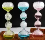 Balançoire en verre coloré avec bulles d'air, composée de trois segments en forme de boule, chacun rempli de liquide coloré (jaune, bleu, rose) et de bulles d'air, sur fond noir.