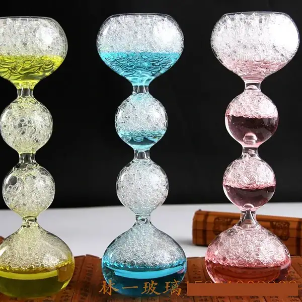 Balançoire en verre coloré avec bulles d'air, composée de trois segments en forme de boule, chacun rempli de liquide coloré (jaune, bleu, rose) et de bulles d'air, sur fond noir.