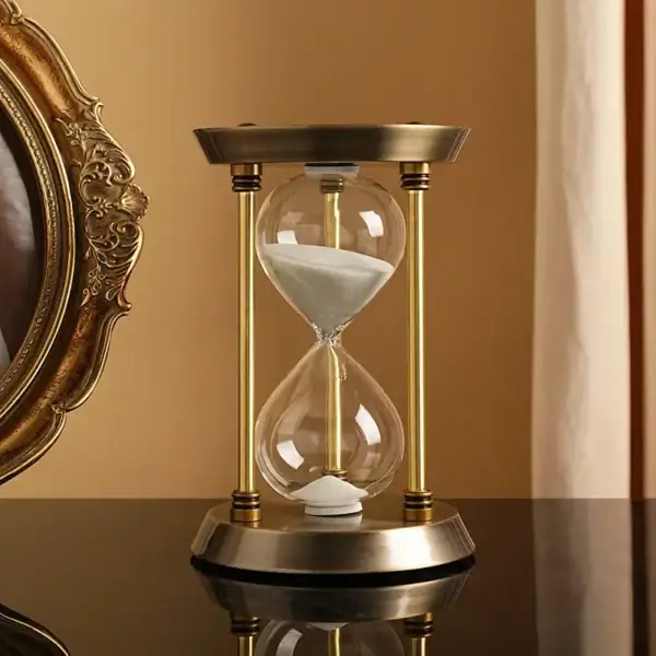 Sablier décoratif en verre avec sable blanc, encadré par une structure en métal doré, posé sur une surface en bois foncé, dans un intérieur élégant.