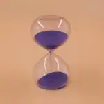 Sablier en verre transparent avec sable violet, symbole du temps qui passe, posé sur un fond beige.