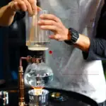 Pipette en verre manipulée par une main lors d'une expérience scientifique, avec un ballon de laboratoire contenant un liquide transparent, illustrant la précision et la technique en chimie.