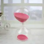 Sablier en verre transparent avec sable rose, symbole du temps qui passe, posé sur une surface claire avec un décor minimaliste en arrière-plan.