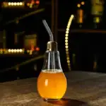 Ampoule décorative en forme de bouteille avec boisson orange, équipée d'une paille métallique, sur une surface en bois dans un environnement sombre et moderne.