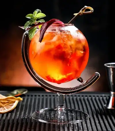 Cocktail coloré dans un verre original avec garniture de menthe et tranche de citron, présenté sur un tapis noir avec accessoires de bar en arrière-plan.