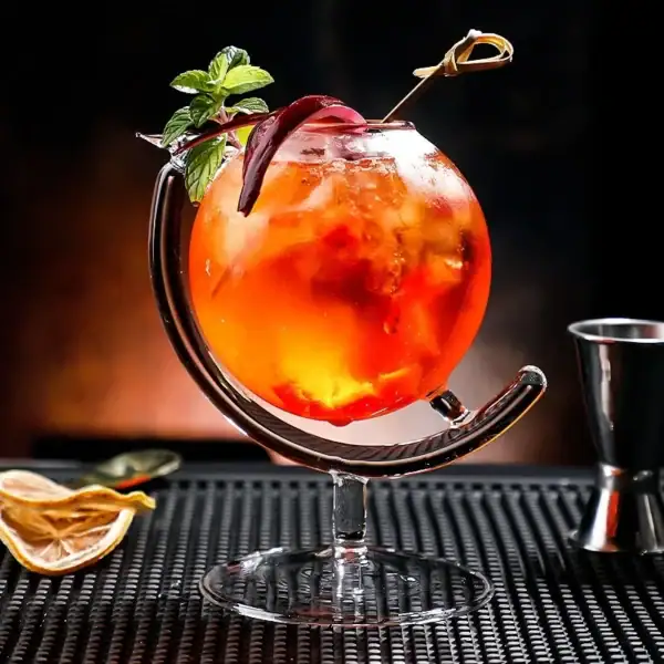 Cocktail coloré dans un verre original avec garniture de menthe et tranche de citron, présenté sur un tapis noir avec accessoires de bar en arrière-plan.