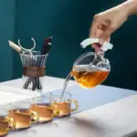 Personne versant du thé chaud dans des tasses en verre, avec une théière en verre à côté, sur une table en bois.
